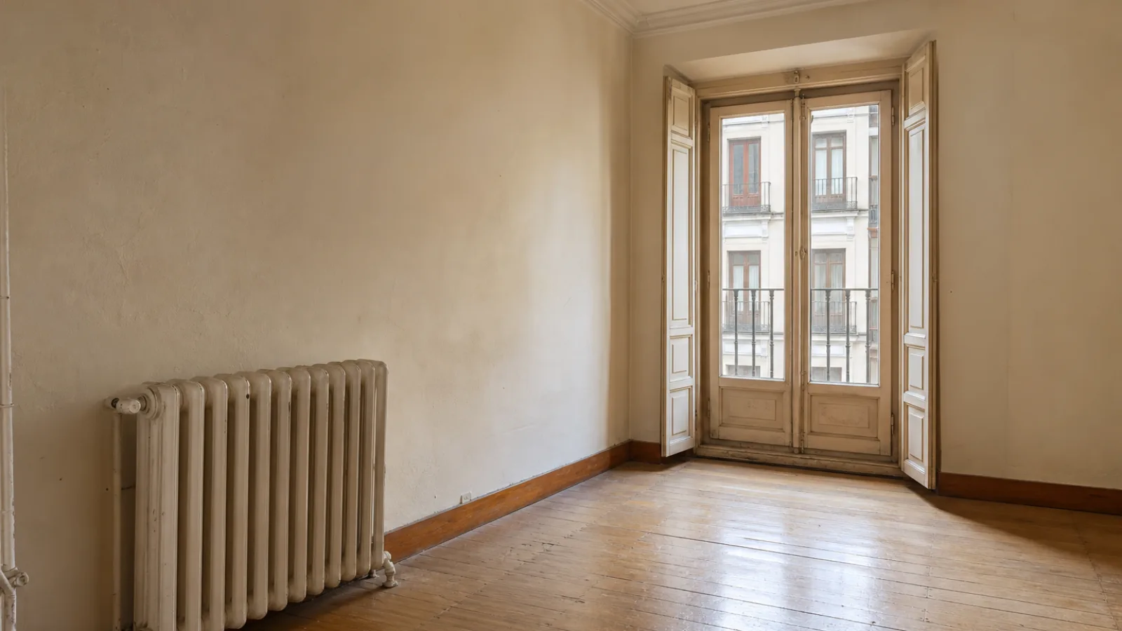 Habitación vacía de un piso antiguo de Madrid con las paredes desnudas, un radiador de hierro fundido apoyado en la pared de la izquierda, tarima de madera gastada y un balcón de dos hojas de carpintería de madera con las contraventanas plegadas a los lados, por el que se ve la fachada clara del edificio de enfrente con sus balcones de hierro