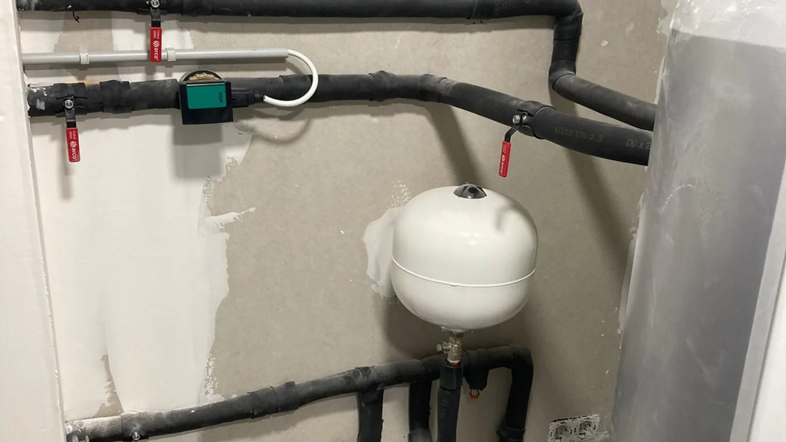 Circuito hidráulico de aerotermia montado en la pared de un cuarto técnico, con tuberías forradas de aislamiento negro, cuatro llaves de corte de maneta roja, la bomba circuladora con su visor, un vaso de expansión blanco colgado en el centro y el lateral del depósito a la derecha