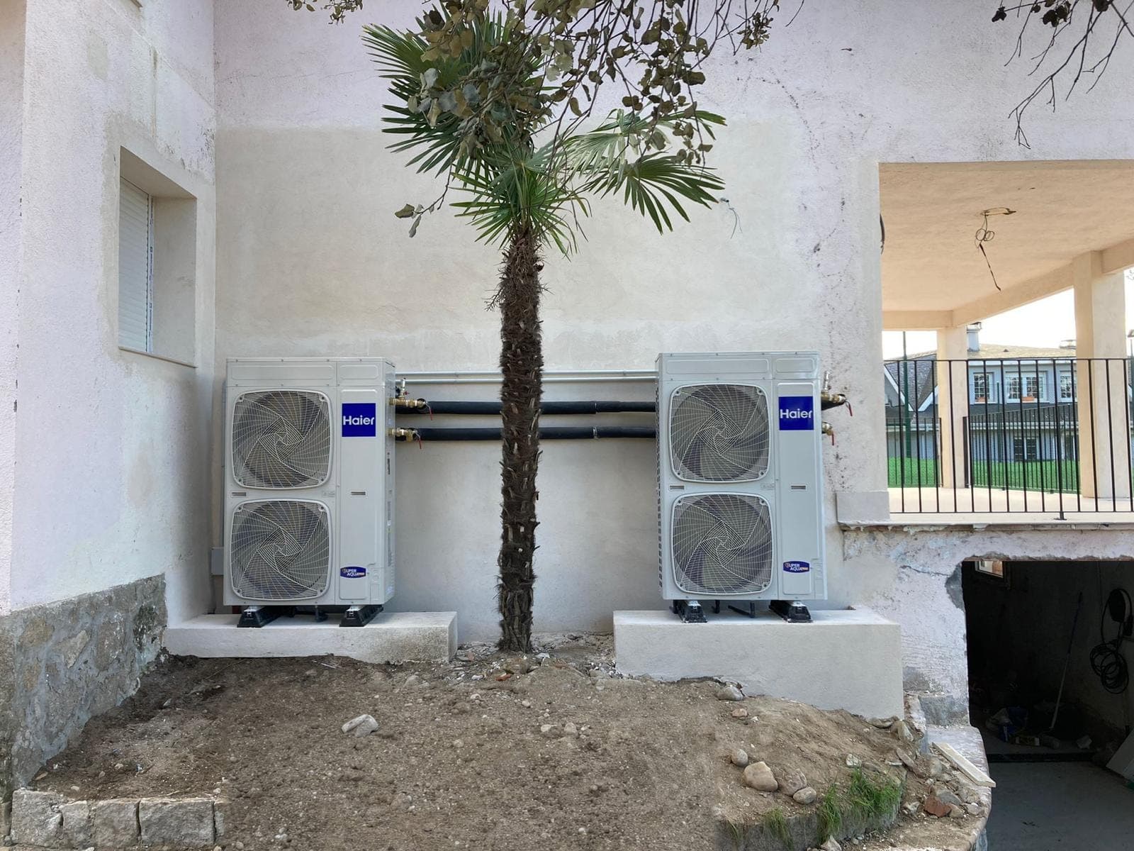 Dos máquinas exteriores de aerotermia Haier instaladas en cascada en el jardín de una vivienda, conectadas por tubería de agua junto a una palmera